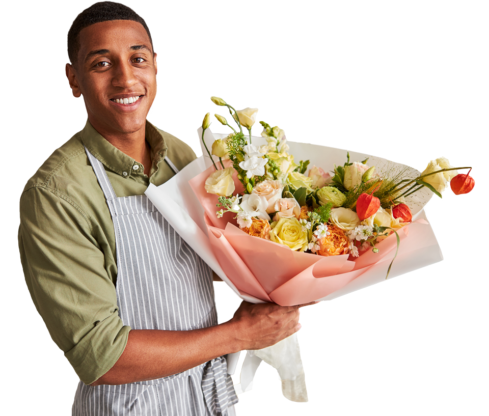 Hombre joven sonriendo, vestido con una camisa verde y un delantal a rayas, sosteniendo un ramo de flores colorido envuelto en papel rosa.