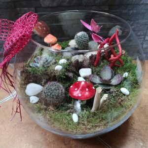 Un terrario en un recipiente de vidrio con decoración de hongos, cactus y suculentas, adornado con elementos como piedras blancas, ramas rojas y una mariposa artificial rosa.