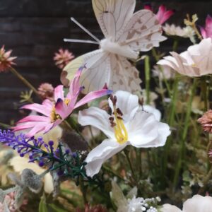 Primer plano de flores silvestres rosadas, blancas y violetas, acompañadas de una mariposa decorativa de tonos beige y marrón.
