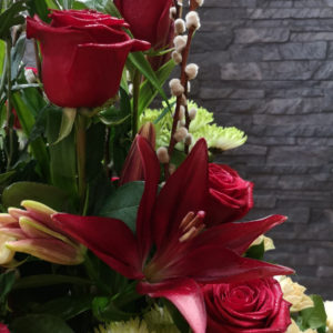 Arreglo floral con lirios rojos en floración, ramas de sauce con capullos blancos, rosas rojas y crisantemos verdes, en un diseño elegante y contemporáneo.