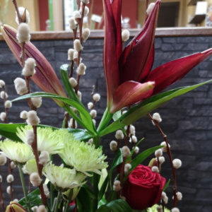 Arreglo floral con lirios rojos en floración, ramas de sauce con capullos blancos, rosas rojas y crisantemos verdes, en un diseño elegante y contemporáneo.