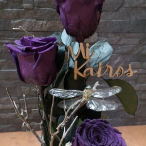 Tres rosas preservadas de color púrpura dispuestas en un arreglo dentro de una base de madera, decoradas con ramas, musgo, piedras, hongos pequeños y una libélula decorativa.
