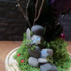 Detalle de la base de un arreglo floral con piedras apiladas, musgo verde, estrellas decorativas, un cristal en forma de obelisco y pequeñas mariquitas rojas.