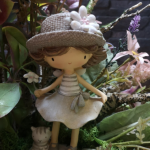 Figura decorativa de una niña con vestido beige y sombrero adornado con flores blancas, rodeada de follaje y flores en un entorno rústico.