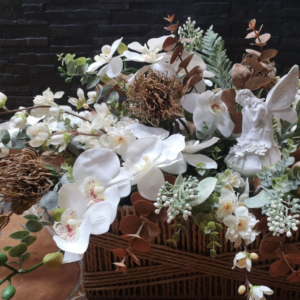 Arreglo floral en cesta decorativa con orquídeas blancas, ramas verdes, hojas marrones, elementos naturales secos y una figura de hada blanca con alas brillantes.