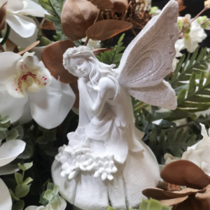 Figura decorativa de un hada blanca con alas brillantes, rodeada de orquídeas blancas, follaje verde y hojas marrones en un ambiente natural y elegante.