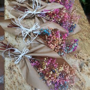 Varios ramos de flores secas teñidas en colores vibrantes, envueltos en papel kraft y atados con lazos blancos, dispuestos sobre una superficie de mármol.
